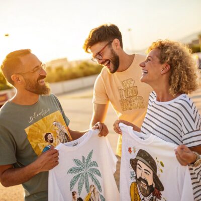 Three friends laughing and holding T-shirts with colorful artistic designs, standing outdoors in bright, golden sunlight.