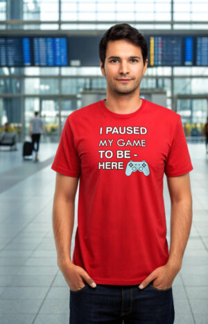 Young man in a red T-shirt with the text “I Paused My Game to Be Here” and a game controller icon, standing in an airport terminal.