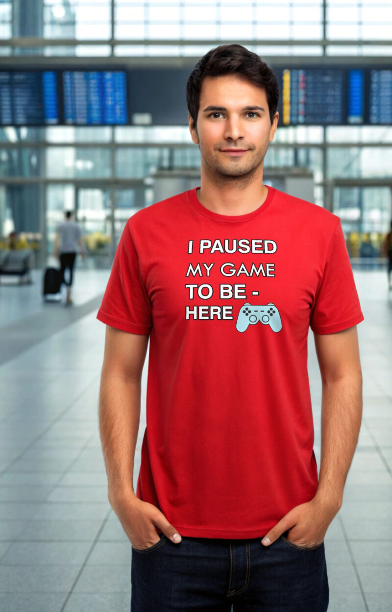 Young man in a red T-shirt with the text “I Paused My Game to Be Here” and a game controller icon, standing in an airport terminal.