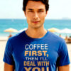 Young man in a blue T-shirt with the text “Coffee First, Then I’ll Deal With You,” standing on a beach.