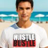 Young man in a white T-shirt with bold “Hustle & Bustle” typography design, standing on a sunny beach.