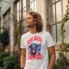 Confident man in a white T-shirt with a bull illustration and American flag, featuring the text “Ride Hard, Stand Proud,” standing near a vintage building with large windows.