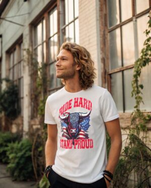 Confident man in a white T-shirt with a bull illustration and American flag, featuring the text “Ride Hard, Stand Proud,” standing near a vintage building with large windows.