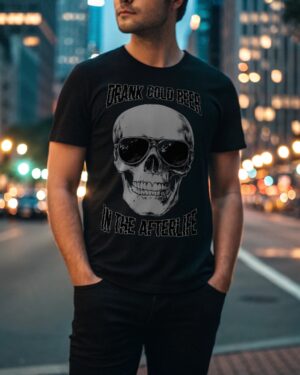Man wearing a black T-shirt with a skull in sunglasses and the phrase “Drink Cold Beer in the Afterlife,” standing on a city street at night.