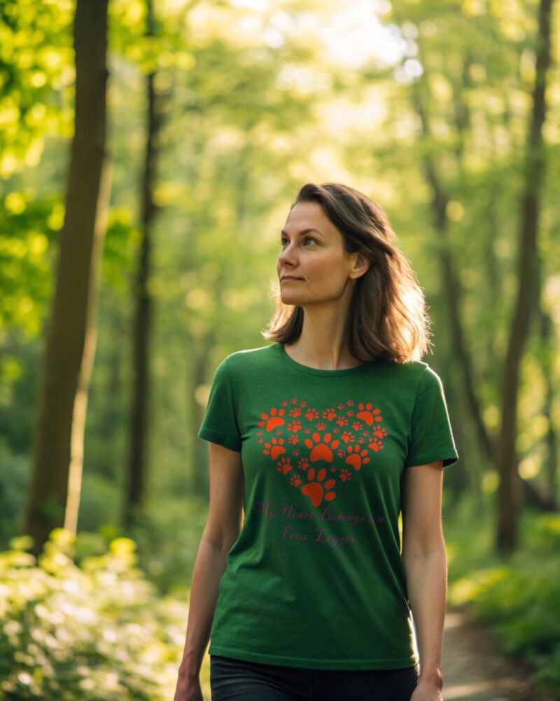 Woman in a green T-shirt with a red paw print heart design and the phrase “My Heart Belongs to a Four Legger,” walking in a sunlit forest.