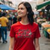 Smiling young woman in a red T-shirt with the quote “Drink Coffee and Smile,” walking through a vibrant outdoor market.