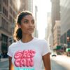 Confident woman in a white T-shirt with bold pink “Girls Can” text and “Yes She Can” slogan, walking through a city street.