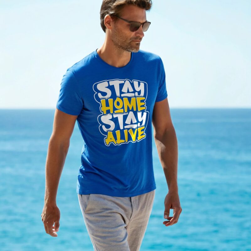 A man in a blue T-shirt with the bold slogan "Stay Home Stay Alive" printed in white and yellow letters