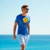 Man wearing a blue T-shirt with a "Sleep Less, Dream More" print, walking by the seaside on a sunny day.