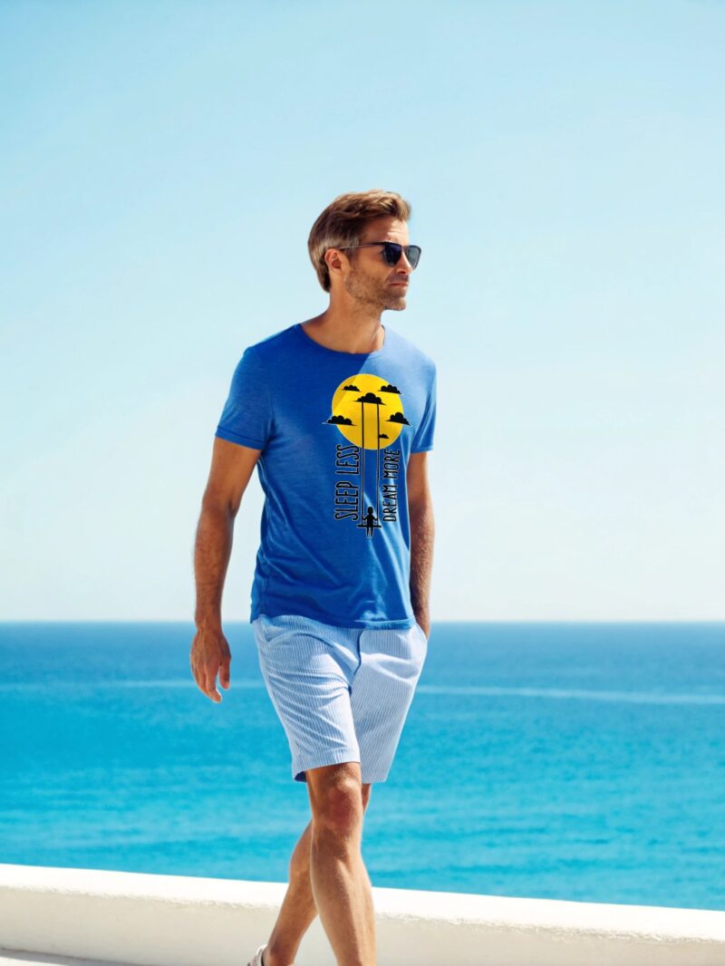 Man wearing a blue T-shirt with a "Sleep Less, Dream More" print, walking by the seaside on a sunny day.