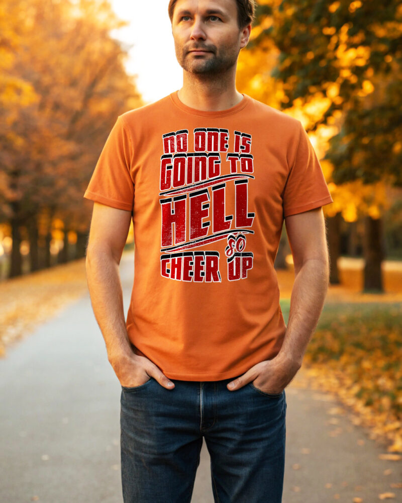 Man wearing an orange T-shirt with bold red and white text "No one is going to Hell — so cheer up", standing on a park path surrounded by autumn trees.