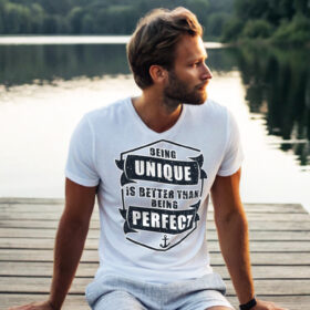 Man in white T-shirt with the phrase "Being Unique Is Better Than Being Perfect" sitting on a dock by a lake.