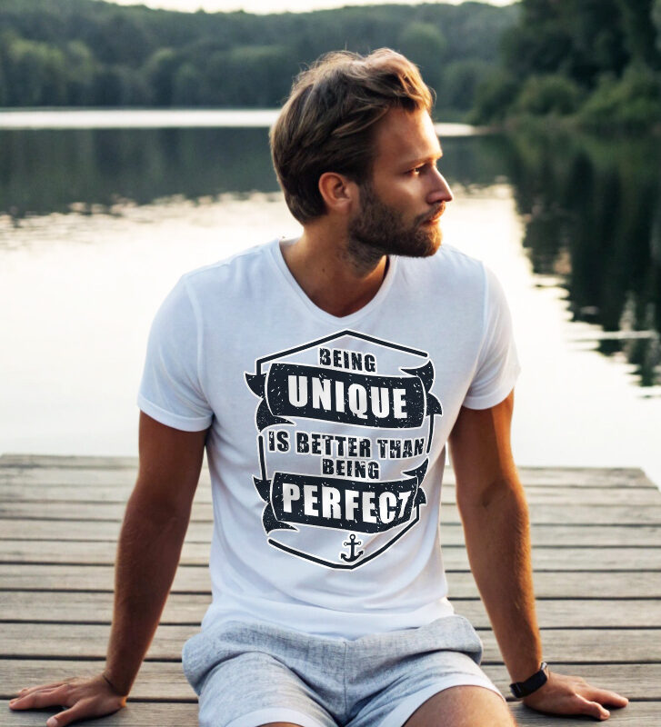 Man in white T-shirt with the phrase "Being Unique Is Better Than Being Perfect" sitting on a dock by a lake.