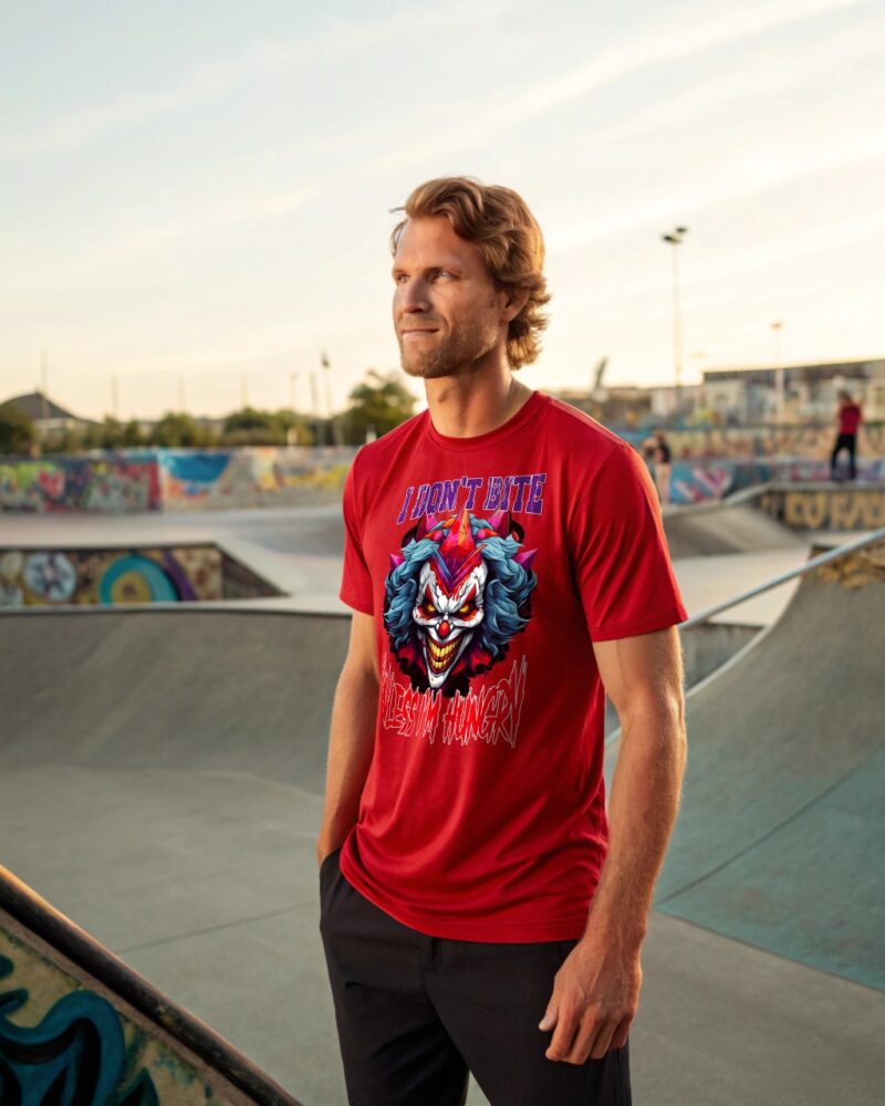 Man in a red T-shirt with a scary clown graphic and the text “I Don’t Bite Unless I’m Hungry,” standing in a graffiti-covered skate park at sunset.