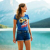 Smiling woman wearing a blue T-shirt with sunflowers, American flag, and the word “Freedom,” standing near a scenic lake with mountains in the background.