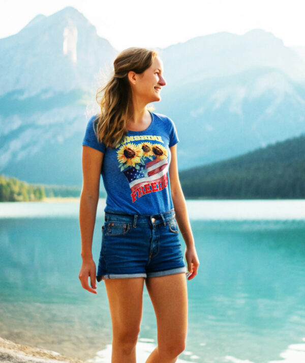 Smiling woman wearing a blue T-shirt with sunflowers, American flag, and the word “Freedom,” standing near a scenic lake with mountains in the background.