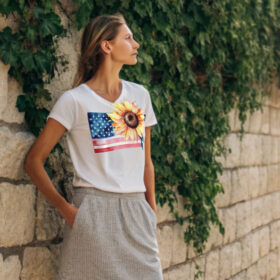 Woman wearing a white T-shirt with a sunflower and American flag print, standing by a stone wall covered in ivy.