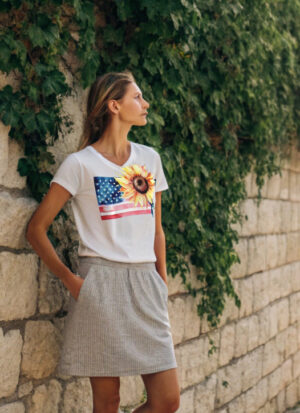 Woman wearing a white T-shirt with a sunflower and American flag print, standing by a stone wall covered in ivy.
