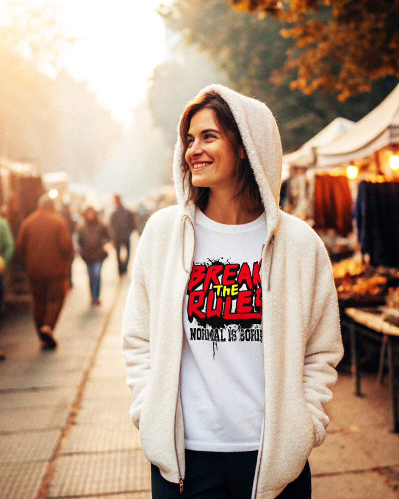Smiling woman wearing a white T-shirt with bold red and black graffiti-style text "Break the Rules – Normal is Boring", walking through a cozy outdoor market in autumn.