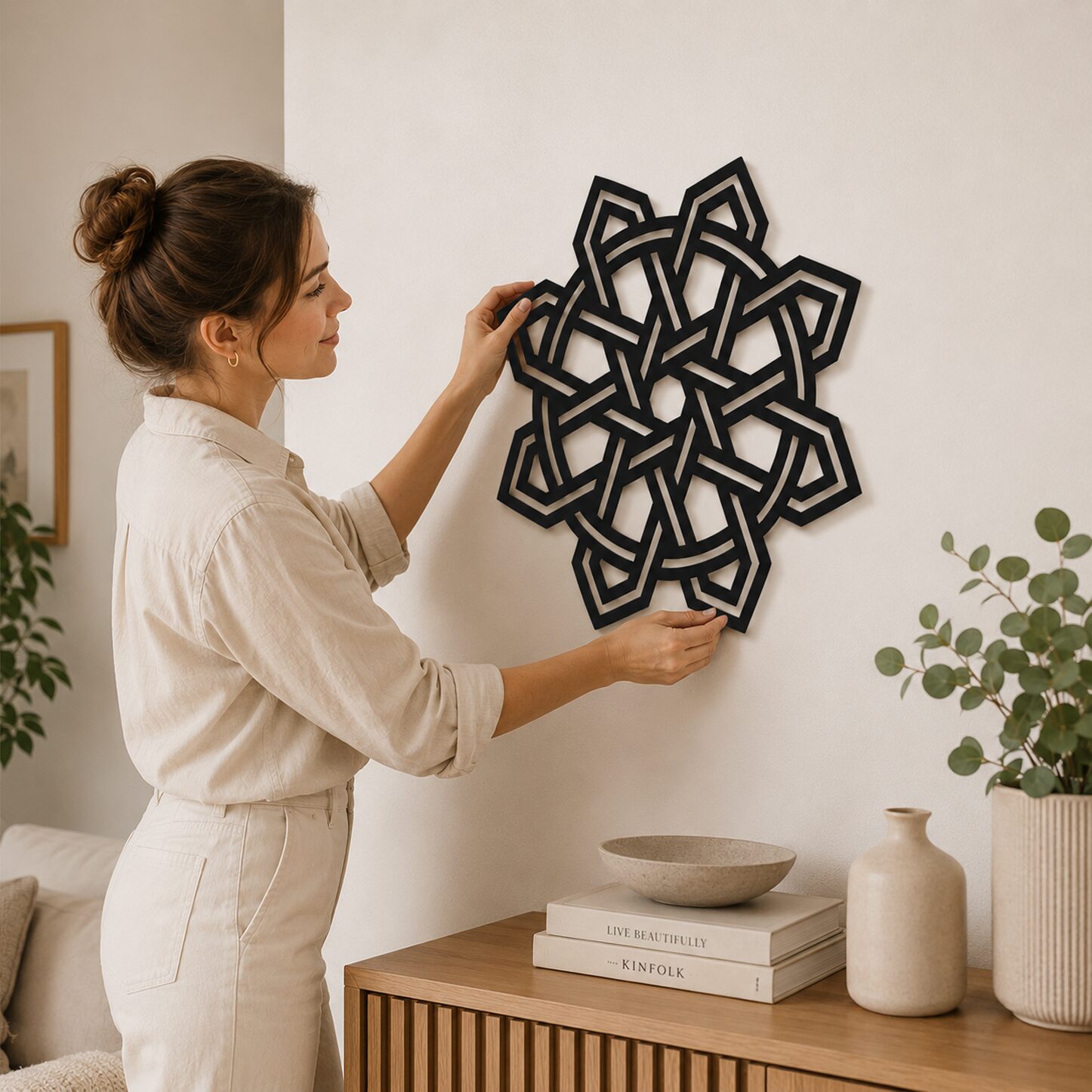 Woman positioning black Celtic Knot wall decor on a neutral wall above a wooden console in a modern interior.