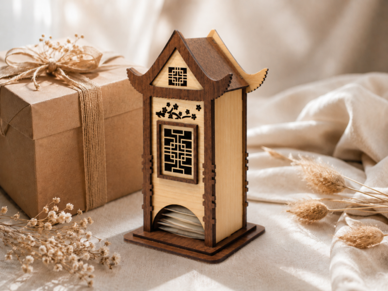 Wooden tea bag organizer shaped like a decorative house, displayed beside a wrapped gift with tea bags visible through the bottom opening.